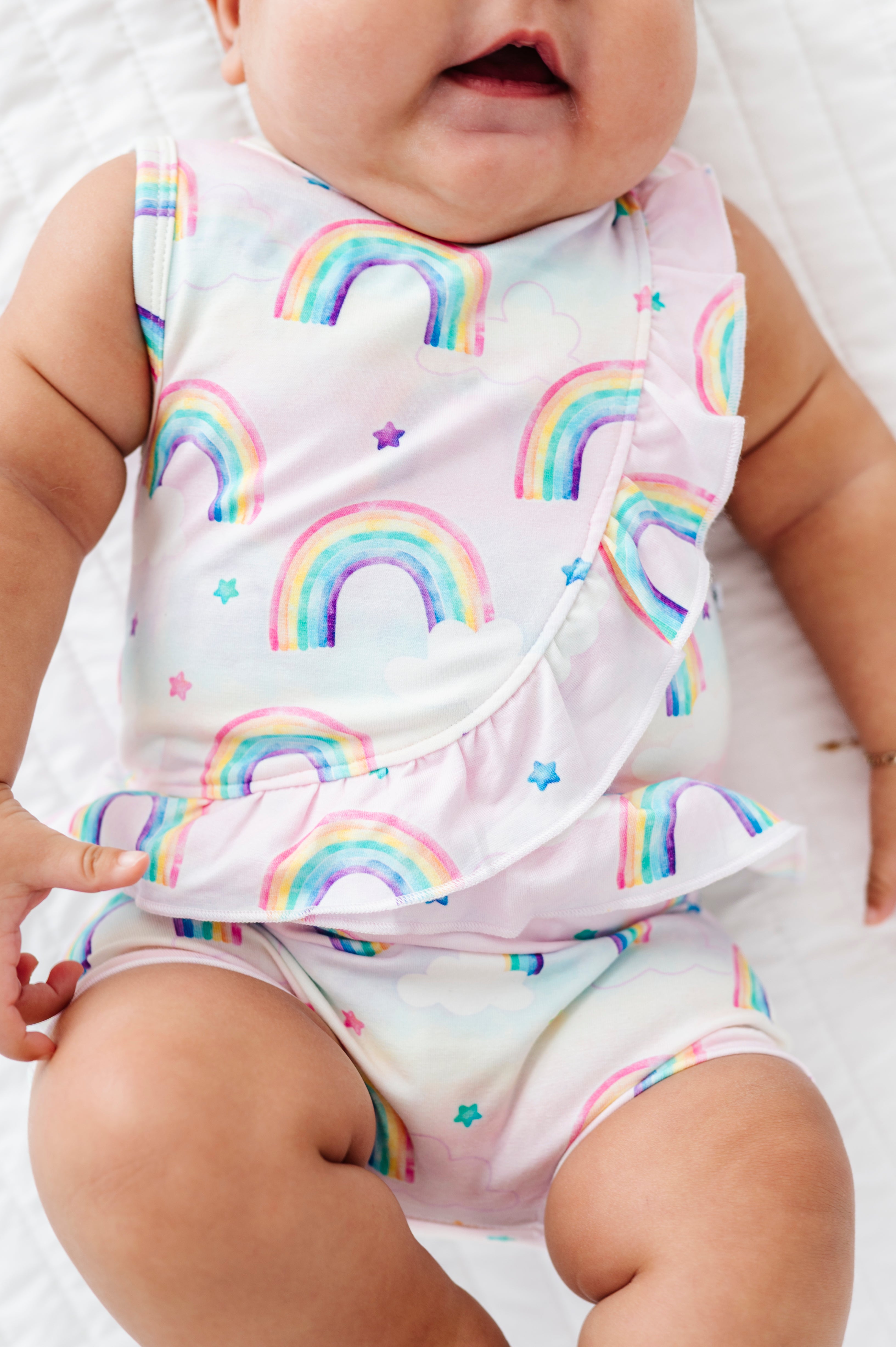 Baby wearing a sleeveless bamboo cotton peplum top and bloomers with watercolor rainbow patterns on a plain background