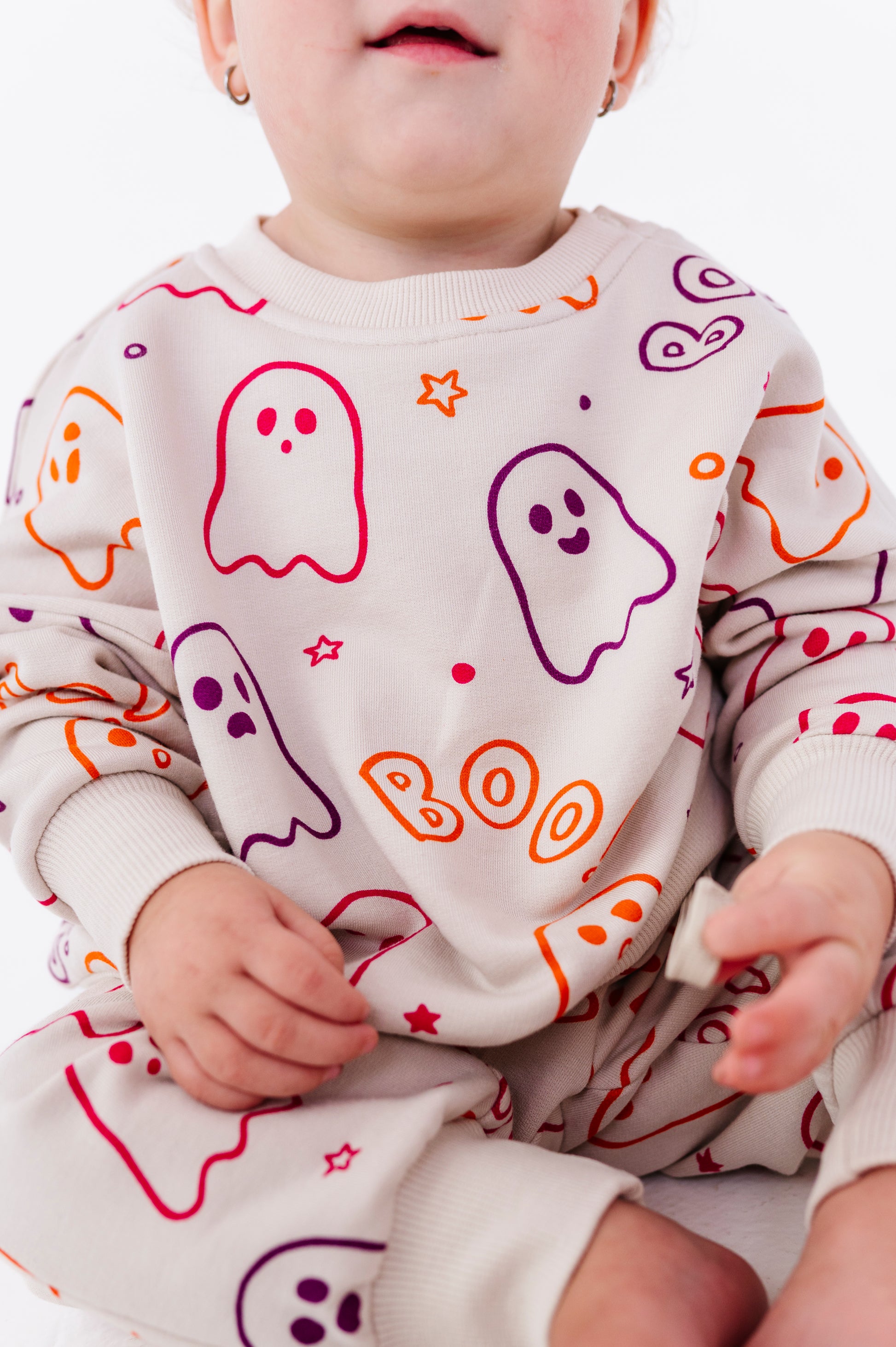 A baby wearing a cream-colored long-sleeve sweatshirt and matching pants set with a colorful Halloween ghost pattern in orange, purple, and red. The sweatshirt features the word 'BOO' printed in orange.