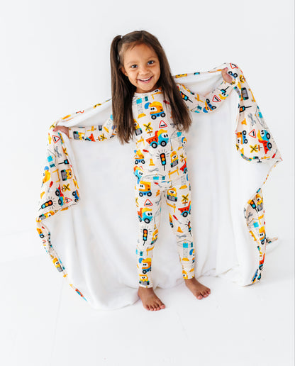 A young girl with long brown hair stands barefoot on a white studio background, holding up a large white blanket or sheet with a colorful construction vehicle print. She is wearing matching printed pajamas featuring toy trucks, cranes, and construction equipment.