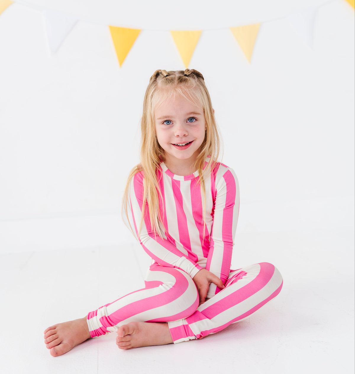 A young girl with blonde hair sitting cross-legged on a white floor, wearing a pink and white vertically striped long-sleeve one-piece jumpsuit. She is posed in a bright studio setting with a yellow and white bunting banner in the background.
