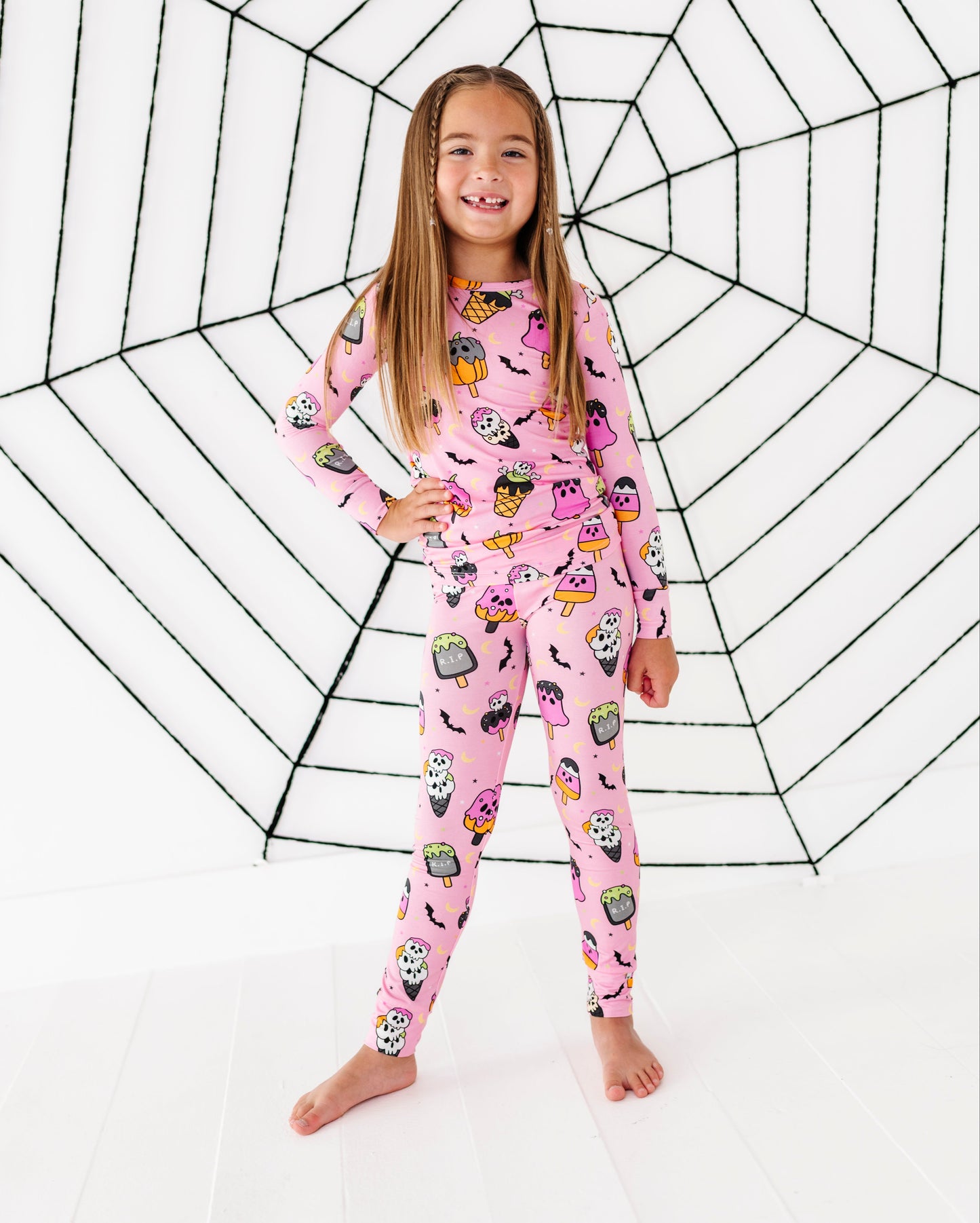 A young girl stands barefoot in front of a large black spiderweb graphic on a white background, wearing a pink long-sleeve pajama set with a colorful cartoon spider-themed print.