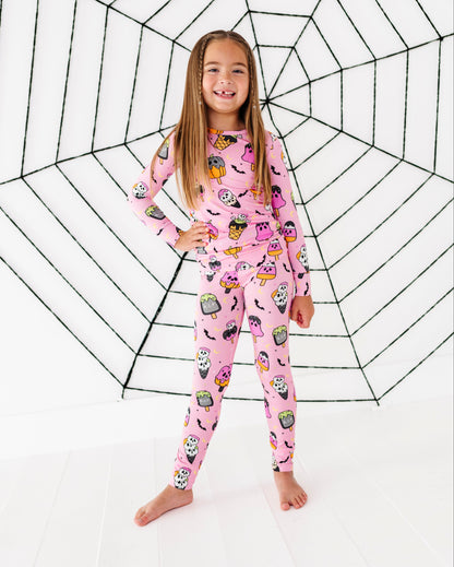 A young girl stands barefoot in front of a large black spiderweb graphic on a white background, wearing a pink long-sleeve pajama set with a colorful cartoon spider-themed print.
