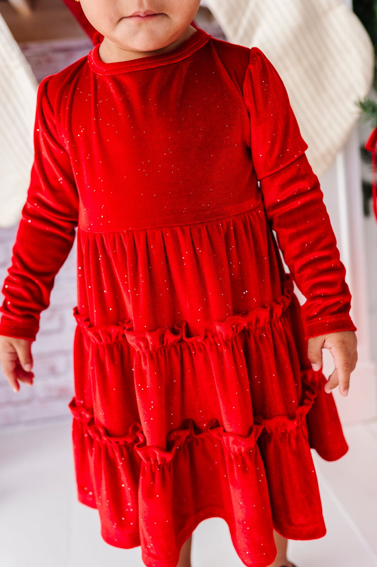 A toddler wearing a bright red long-sleeve dress with a tiered ruffle skirt and subtle glitter, standing in a festive indoor setting.