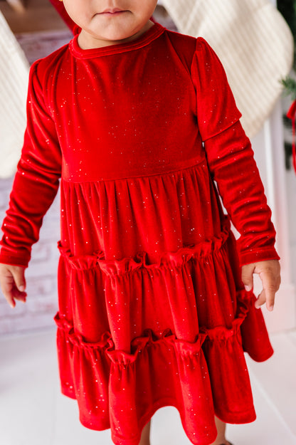 A toddler wearing a bright red long-sleeve dress with a tiered ruffle skirt and subtle glitter, standing in a festive indoor setting.