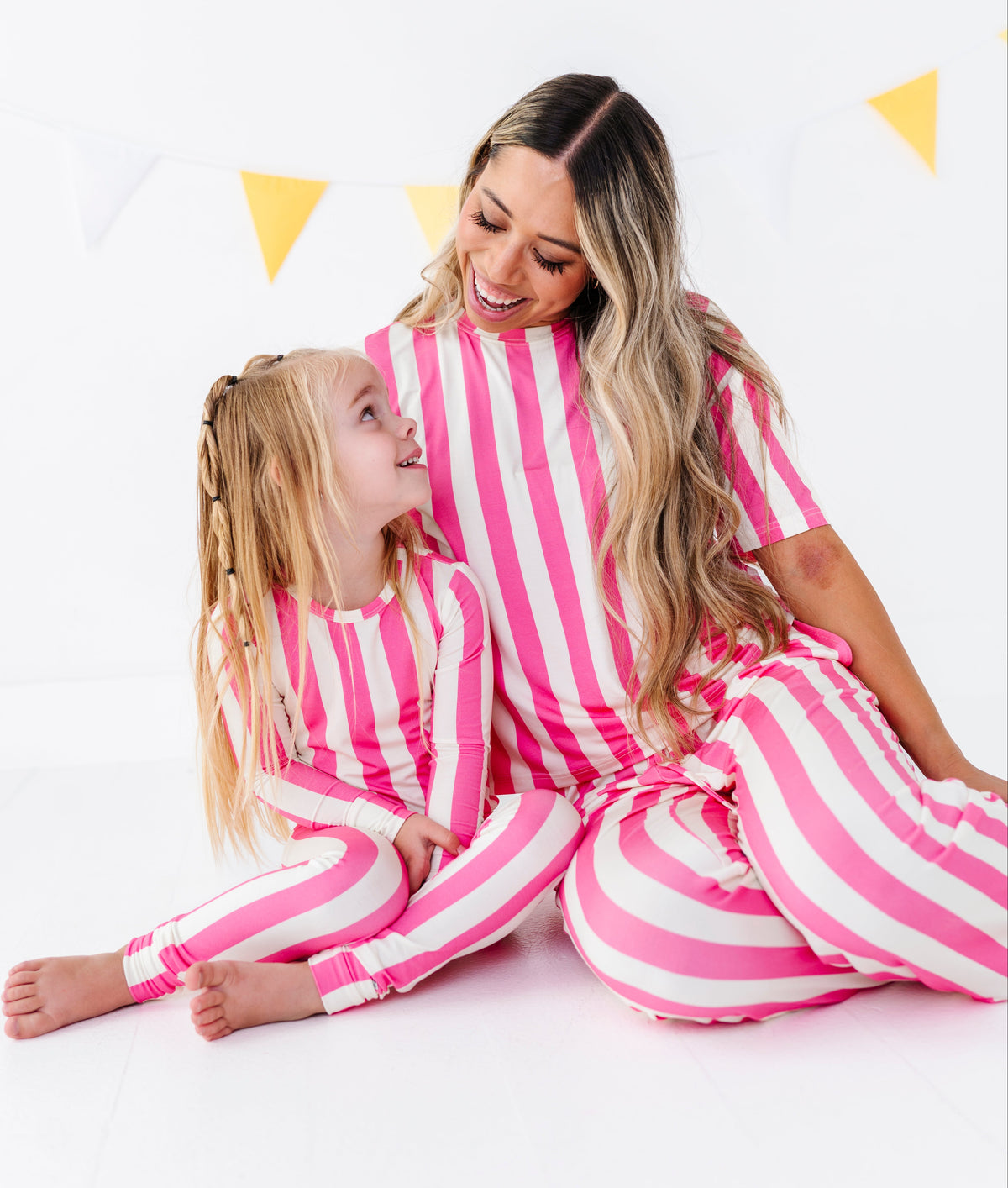 A woman and a young girl sitting together on a white floor, both wearing matching pink and white vertically striped pajamas. The background is a clean white studio setting with a yellow and white bunting banner.