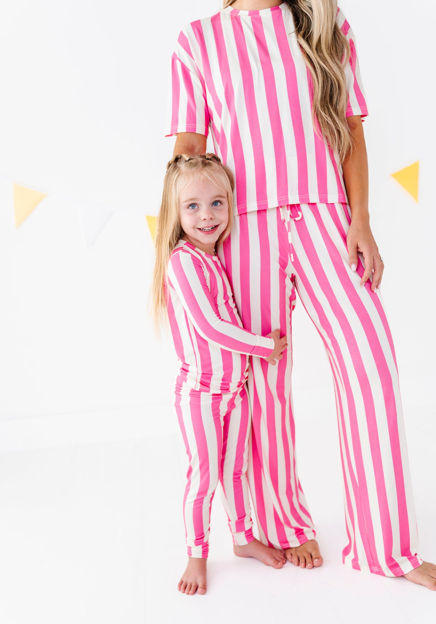 A woman and a young girl wearing matching pink and white vertically striped pajama sets, standing barefoot against a white studio background with subtle yellow triangle decorations.