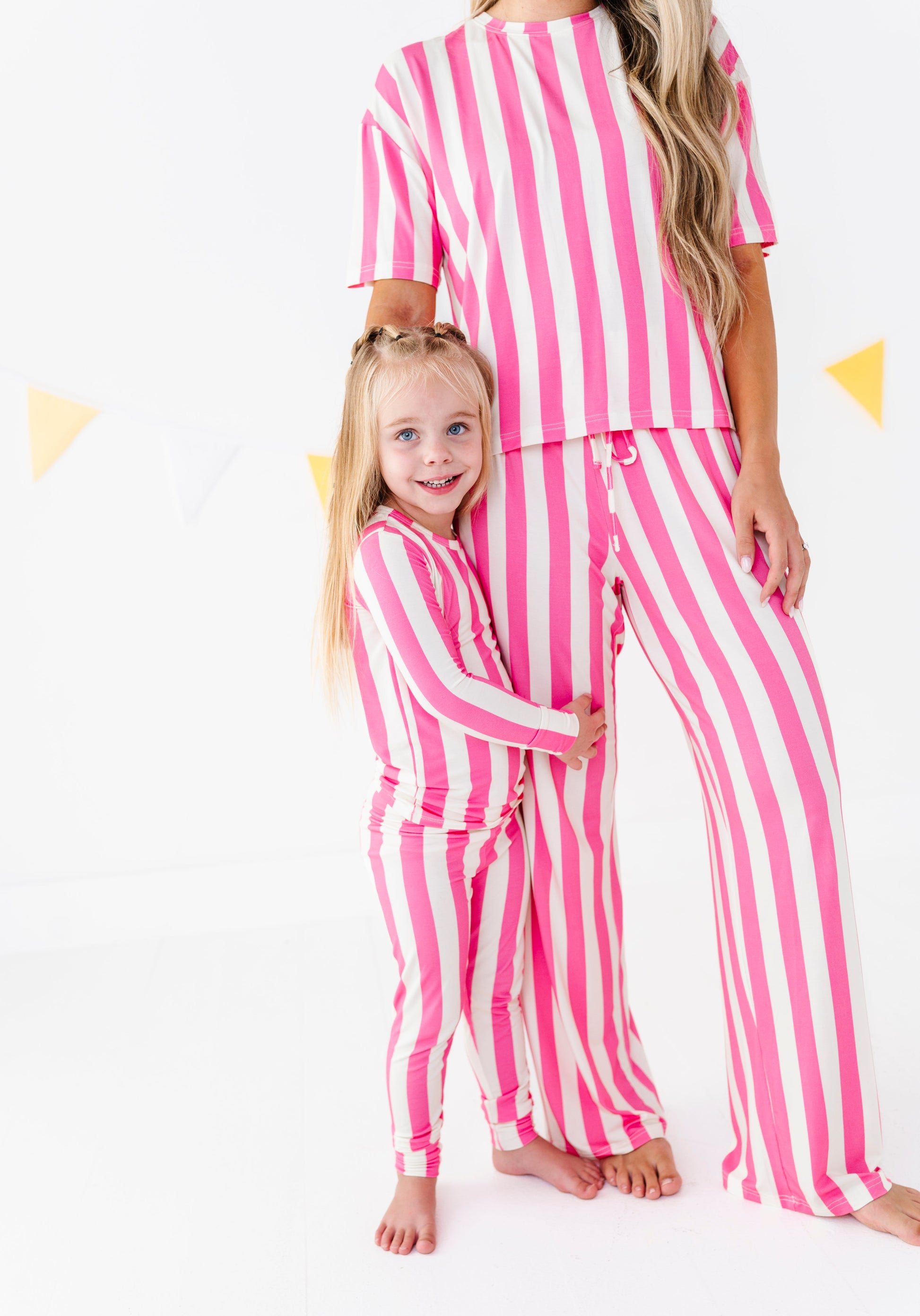 A woman and a young girl wearing matching pink and white vertically striped pajama sets, standing barefoot against a white studio background with subtle yellow triangle decorations.