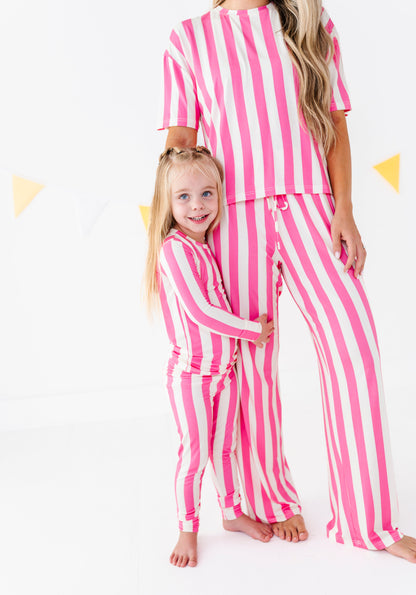 A woman and a young girl wearing matching pink and white vertically striped pajama sets, standing barefoot against a white studio background with subtle yellow triangle decorations.