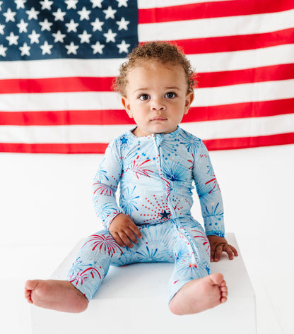 Child wearing a blue bamboo zip romper with red and white firework patterns, sitting in front of an American flag.