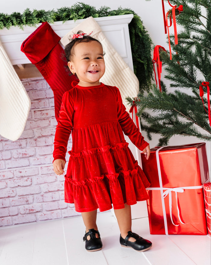 A toddler girl wearing a festive red tiered long-sleeve dress stands in a decorated holiday room next to a Christmas tree and wrapped presents.