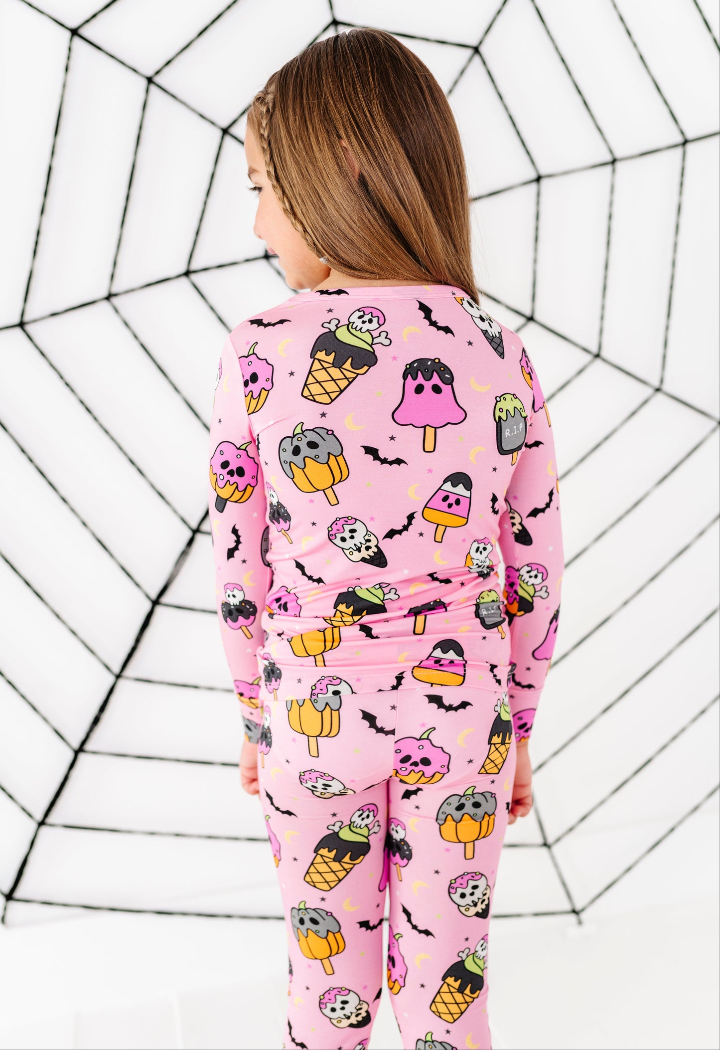 A child wearing a pink long-sleeve pajama set with a Halloween-themed ice cream cone print, shown from the back against a white background with a black spiderweb pattern.