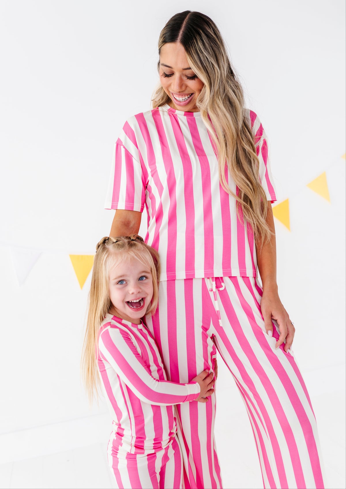 A woman and a young girl wearing matching pink and white vertically striped pajamas, standing against a white background with subtle yellow triangle decorations.