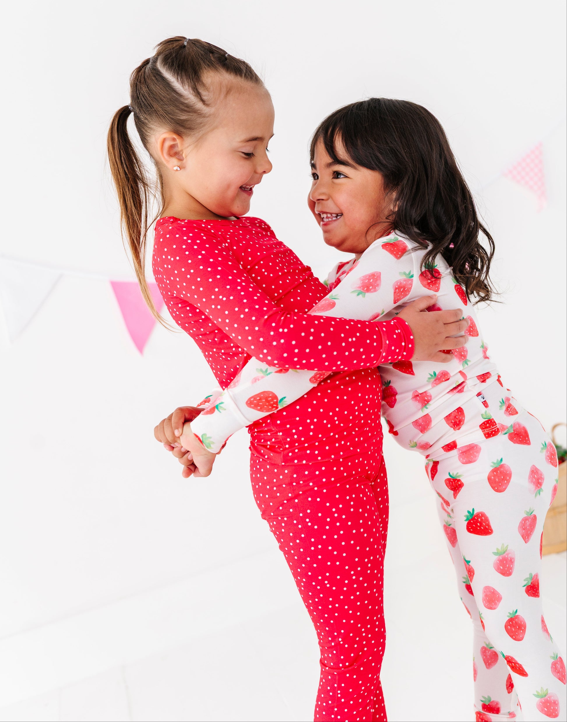 Two children in matching strawberry-themed pajamas hugging on a white background
