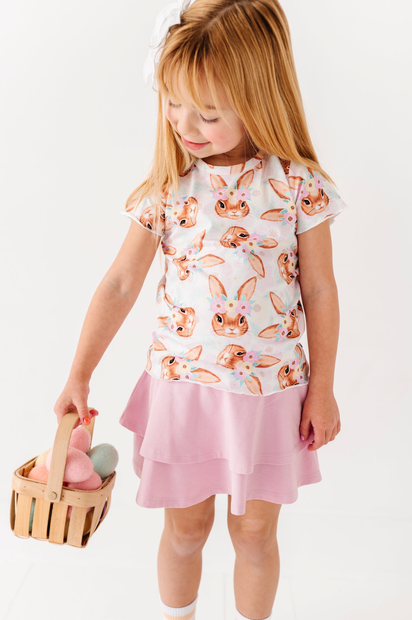 Young girl wearing a bunny print top and pink skirt holding a basket with Easter eggs.
