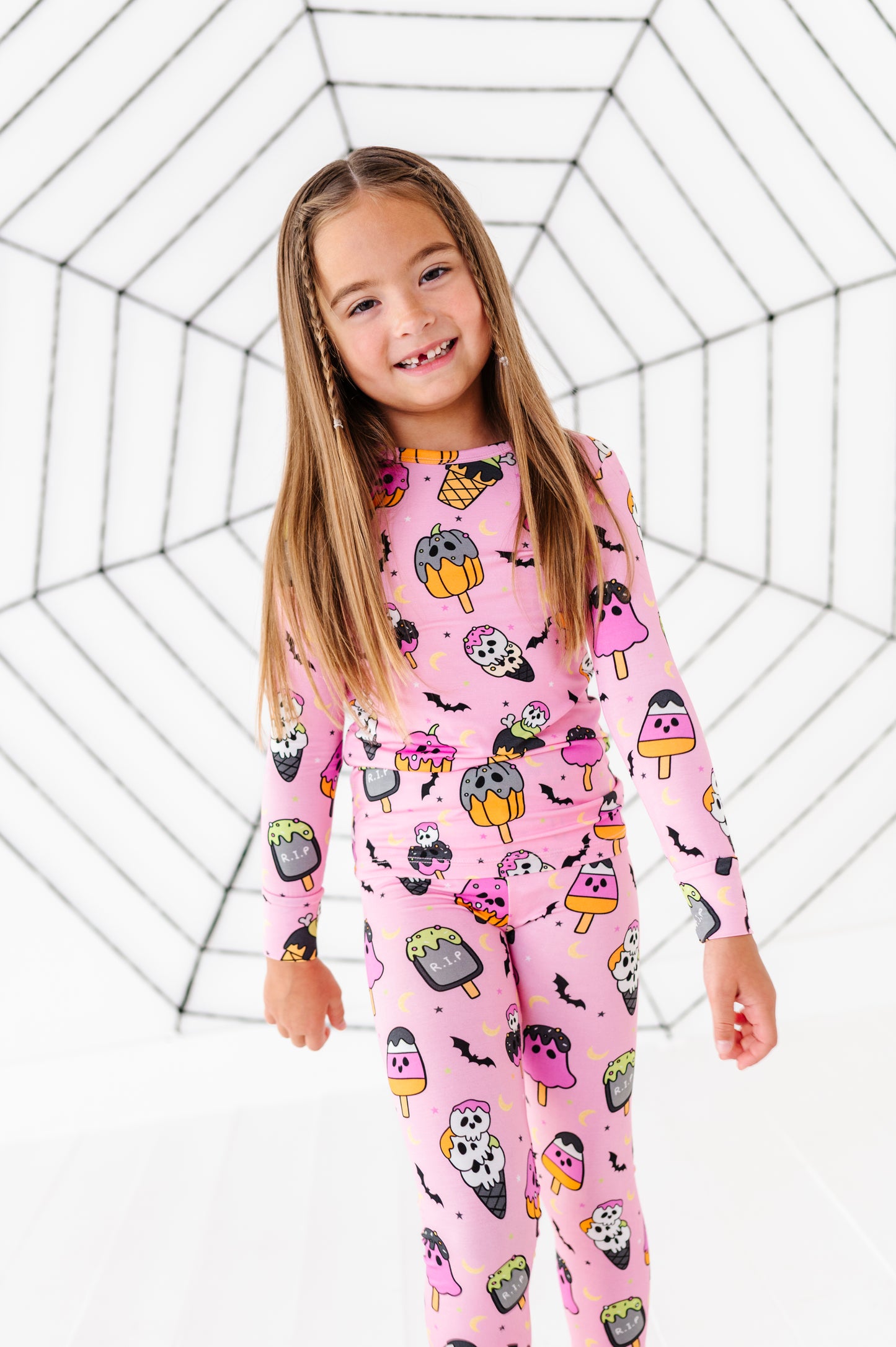 A young girl with long light brown hair stands smiling in a pink long-sleeve pajama set with a cartoon popsicle and bat pattern, posed against a white background with a black spiderweb graphic.