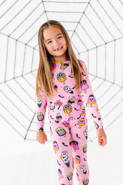 A young girl with long light brown hair stands smiling in a pink long-sleeve pajama set with a cartoon popsicle and bat pattern, posed against a white background with a black spiderweb graphic.