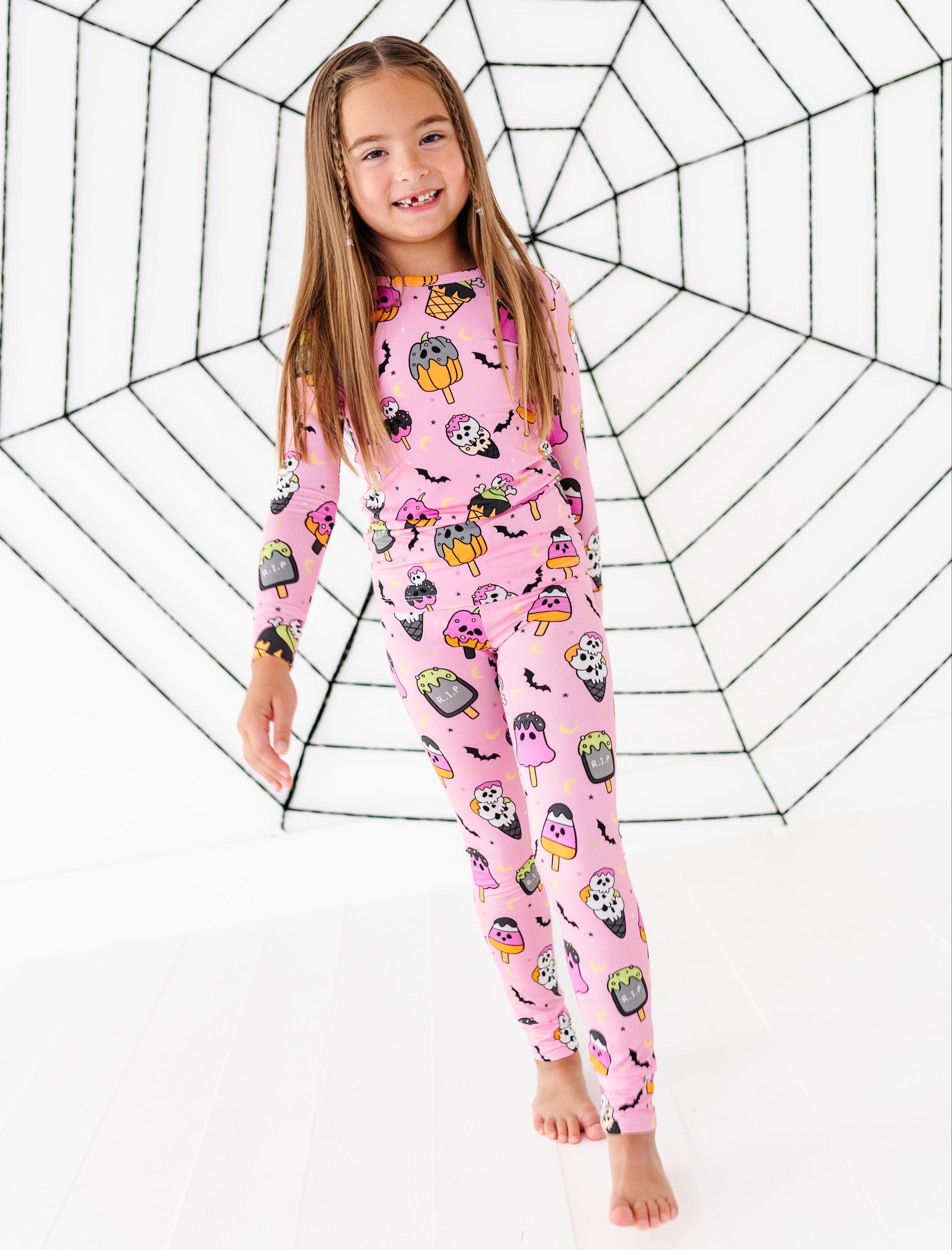 A young girl with long light brown hair stands barefoot in front of a white studio backdrop with a large black spiderweb graphic. She is wearing a pink long-sleeve one-piece pajama set covered in a colorful Halloween-themed print featuring pumpkins, ghosts, skulls, bats, and spiders.