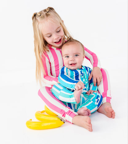 Two children sitting together on a white studio background. The older child has blonde hair and wears a pink, white, and blue striped long-sleeve top and matching bottoms, while the younger child wears a blue and white patterned long-sleeve top and bottoms.