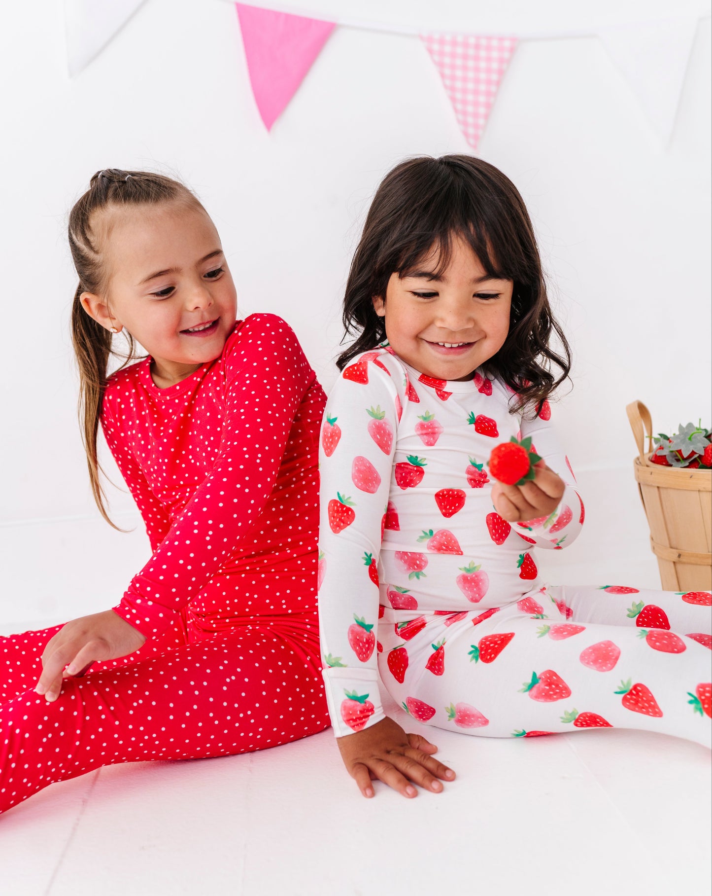 Two children in red and strawberry-patterned pajamas sitting on a bed with a white background.