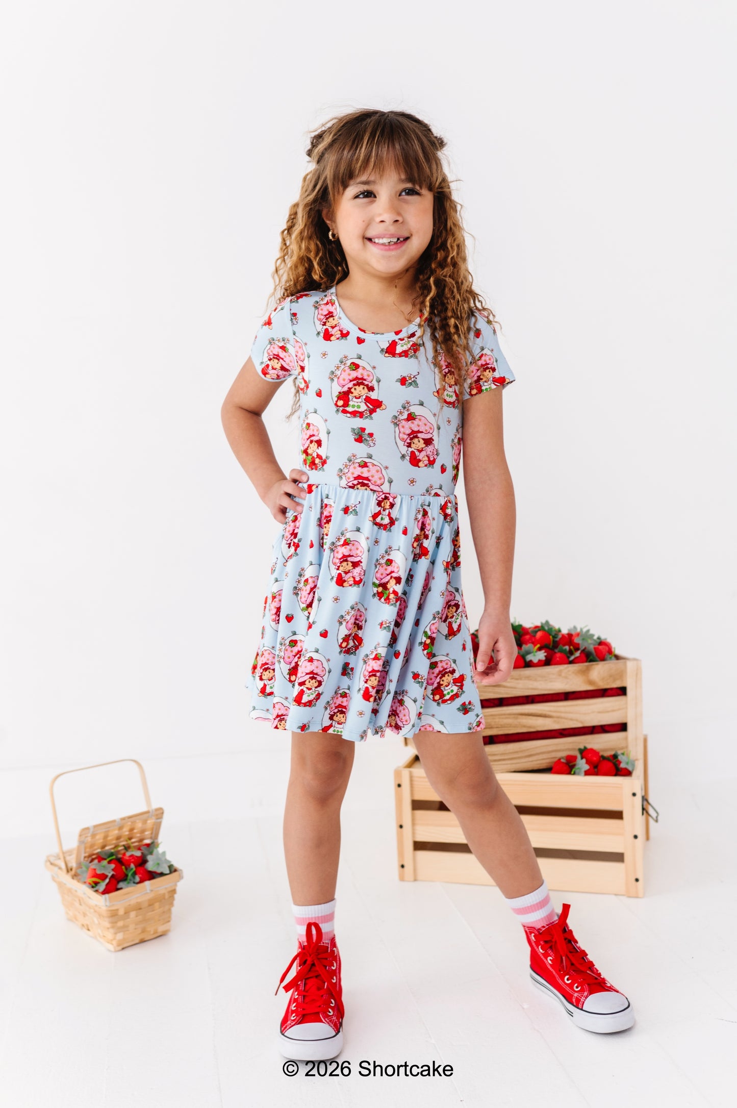 Young girl in a Strawberry Shortcake™ dress with red shoes standing next to wooden crates and baskets on a white background.