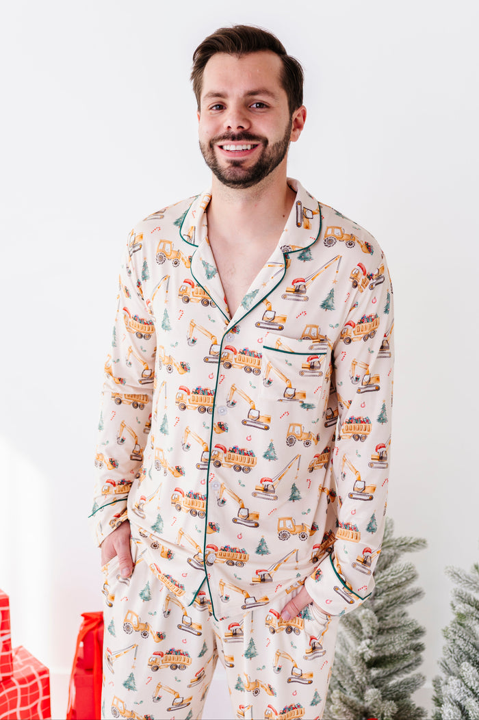 A man wearing a long-sleeve bamboo pajama set with a colorful holiday construction theme print, featuring a cream base with orange, green, and yellow graphics, and dark green piping. He is standing against a white wall with a small Christmas tree and wrapped gifts visible.