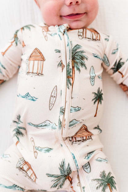 Baby wearing a onesie with beach-themed design, including palm trees and huts.