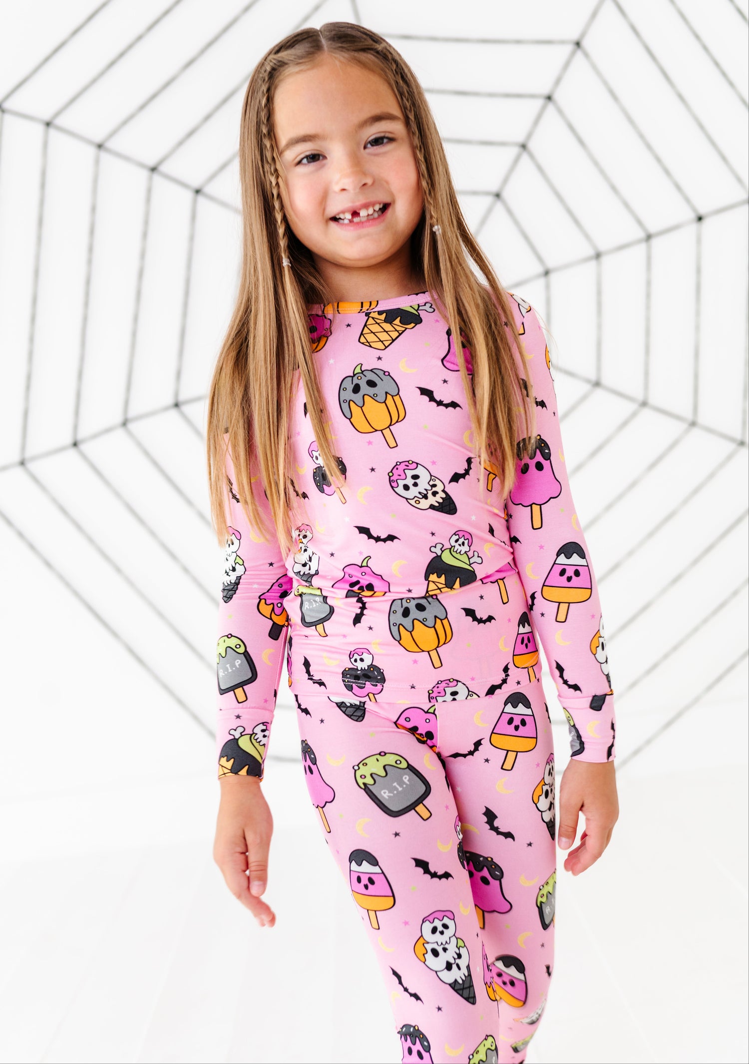 A young girl with long light brown hair stands smiling, wearing a pink long-sleeve pajama set covered in a Halloween-themed print of popsicles, bats, ghosts, and owls. The background is a white studio backdrop with a black spiderweb graphic.