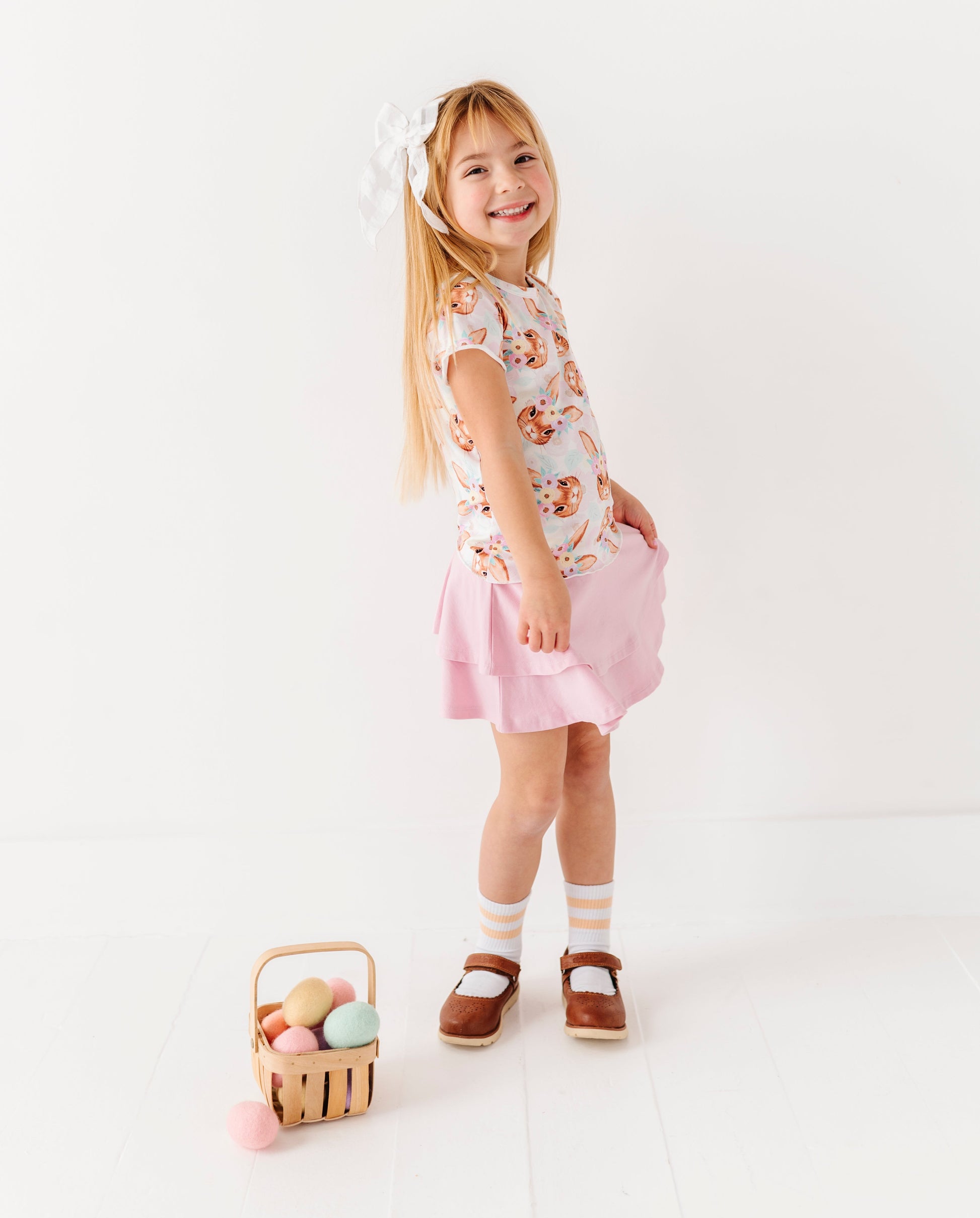 Young girl in a floral top and pink skirt holding a basket of Easter eggs on a white background