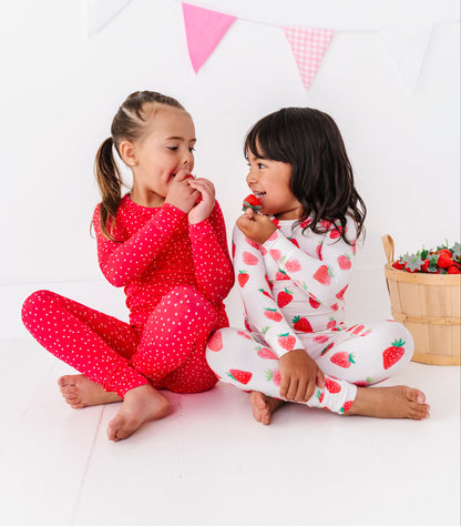 Two children in red and white pajamas sitting on a white floor with pink flags in the background.