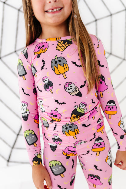 A young girl wearing a pink long-sleeve pajama set with a Halloween ice cream print, featuring popsicles, bats, and skulls, set against a white background with a black spiderweb pattern.