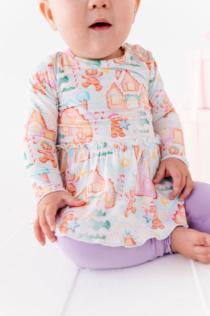A baby girl wearing a Christmas gingerbread village-themed bamboo peplum with light purple bamboo leggings