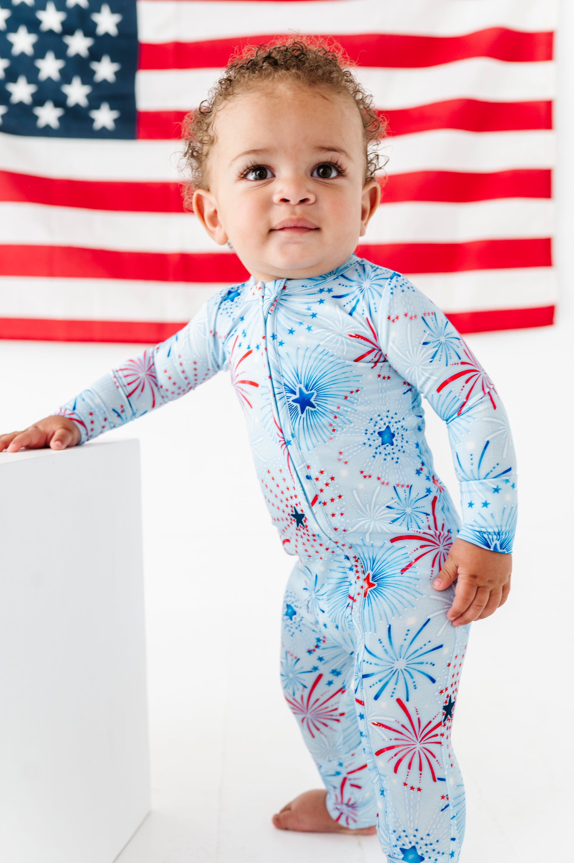Child wearing a patriotic bamboo convertible romper with fireworks design in front of an American flag.