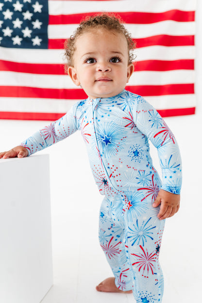 Child wearing a patriotic bamboo convertible romper with fireworks design in front of an American flag.