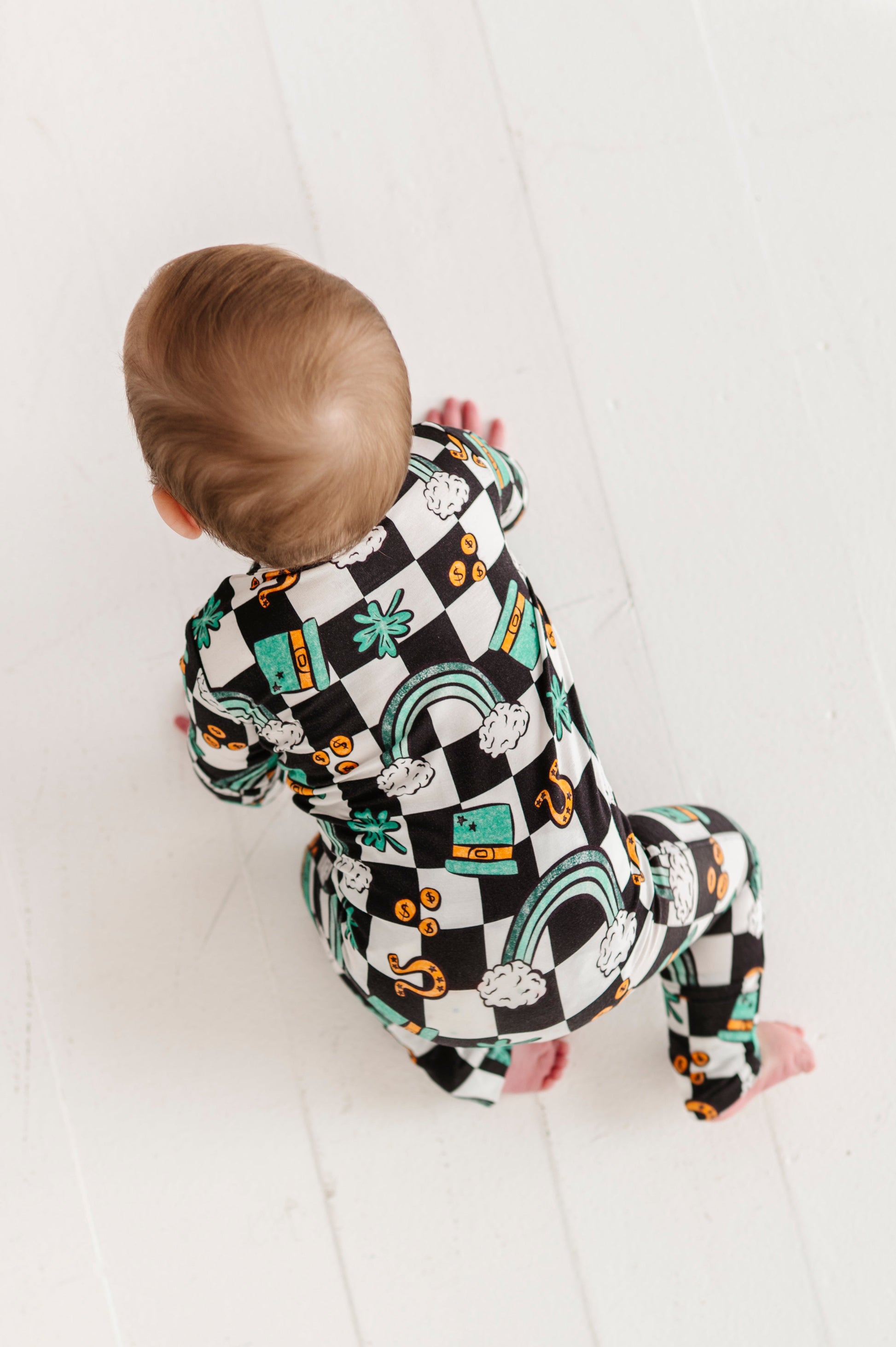Baby crawling on a white surface wearing a bamboo St. Patrick's Day convertible romper featuring green rainbows, top hats, coins, and four leaf clovers 