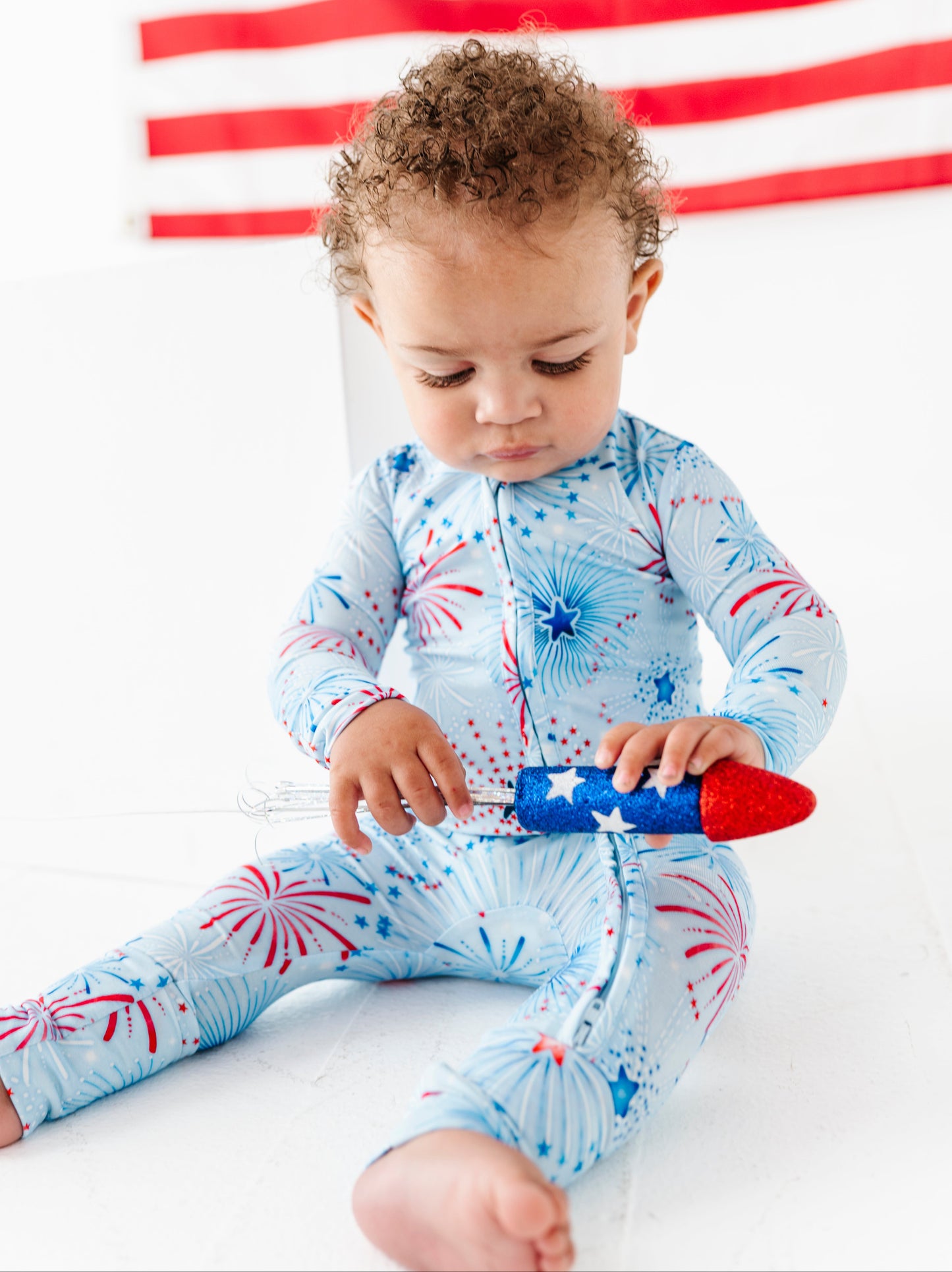 Child wearing a blue bamboo onesie with red and white patterns, sitting on a white surface with an American flag in the background.