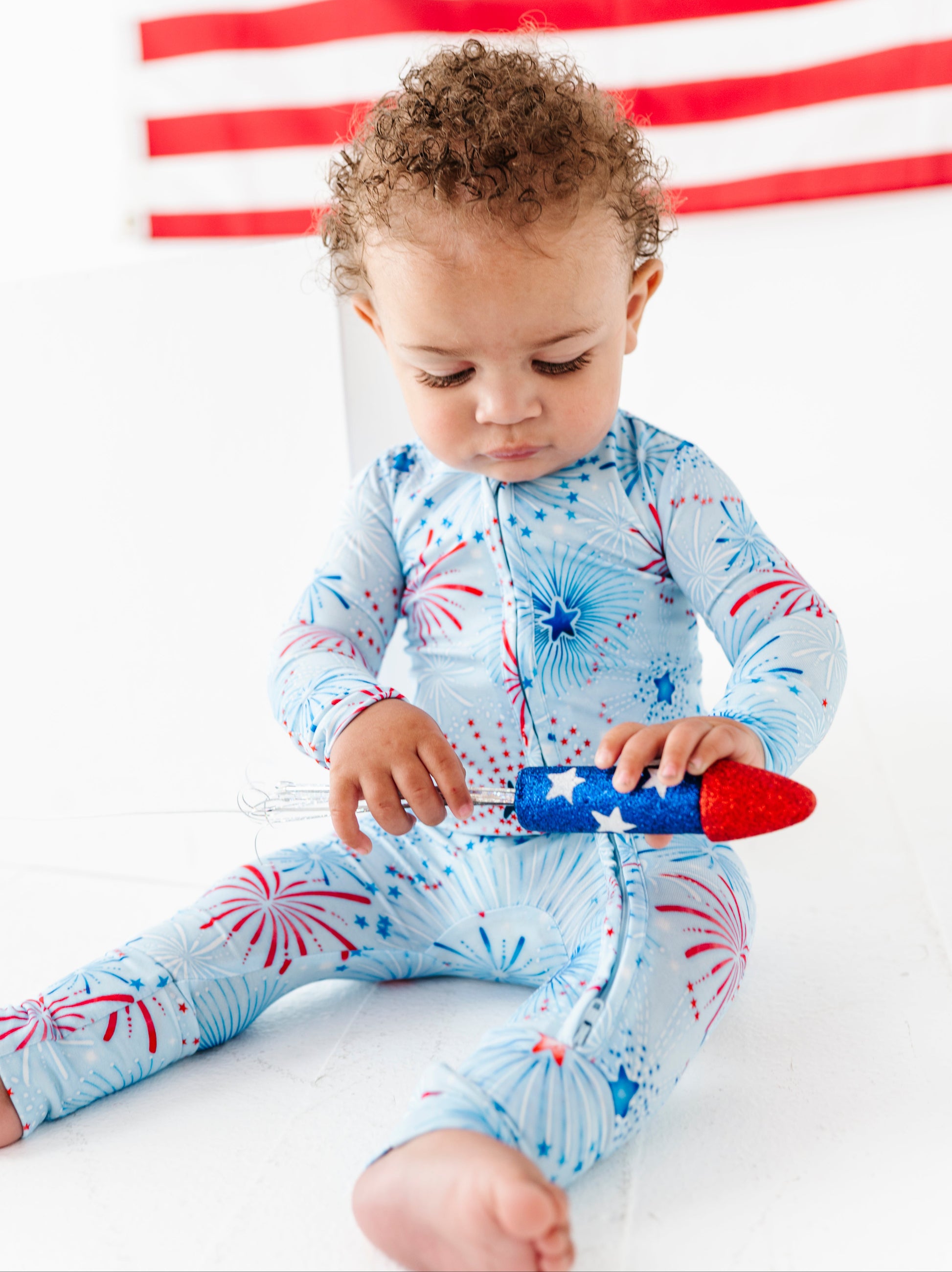 Child wearing a blue bamboo onesie with red and white patterns, sitting on a white surface with an American flag in the background.