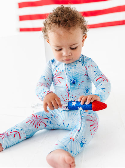 Child wearing a blue bamboo onesie with red and white patterns, sitting on a white surface with an American flag in the background.