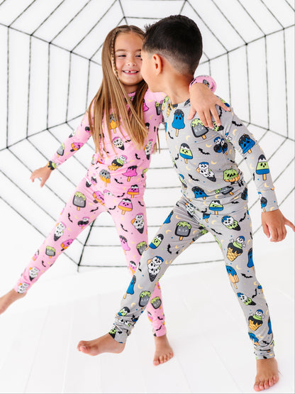 Two children wearing matching Halloween-themed pajama sets pose together in front of a white studio backdrop with a black spiderweb graphic. The girl wears a pink long-sleeve pajama set with a monster print, and the boy wears a gray long-sleeve pajama set with a monster print.