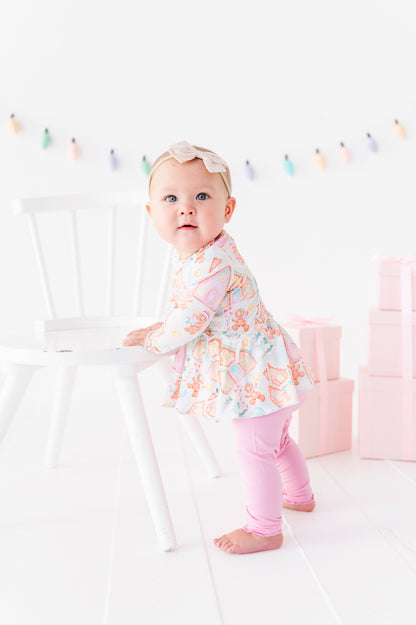 A baby girl wearing a Christmas gingerbread village-themed bamboo peplum with light purple bamboo leggings