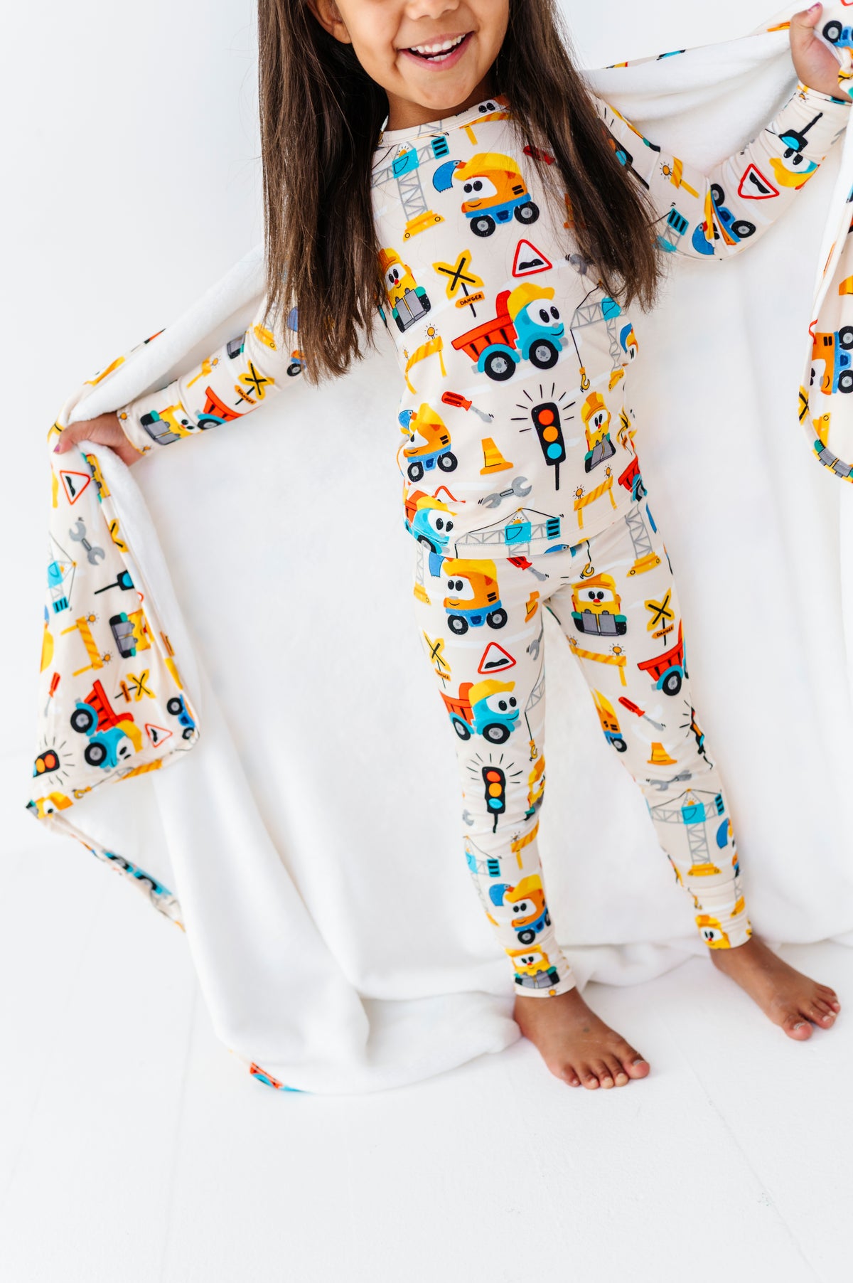 A young girl smiling and holding open a white blanket while wearing a one-piece pajama top and matching pants with a colorful construction vehicle print on a white background.