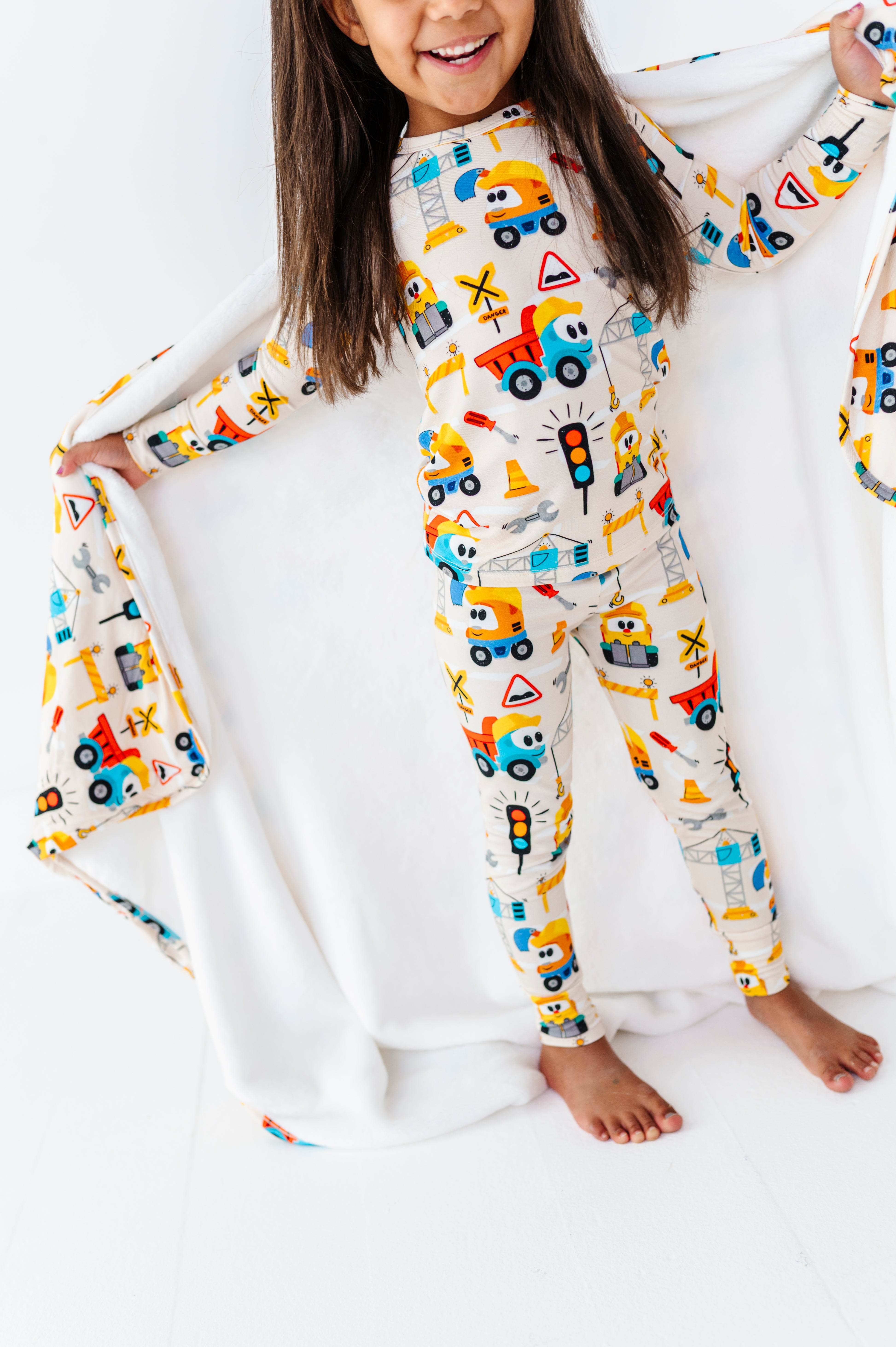 A young girl smiling and holding open a white blanket while wearing a one-piece pajama top and matching pants with a colorful construction vehicle print on a white background.