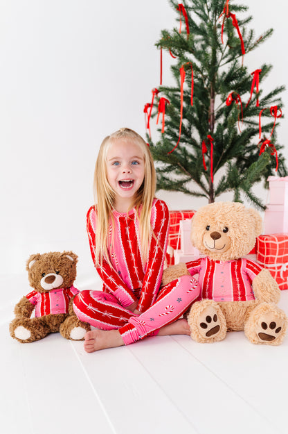 A young girl with blonde hair sits cross-legged on a white floor, wearing a pink and red striped bamboo pajama set. She is surrounded by two teddy bears and wrapped presents in front of a decorated Christmas tree.