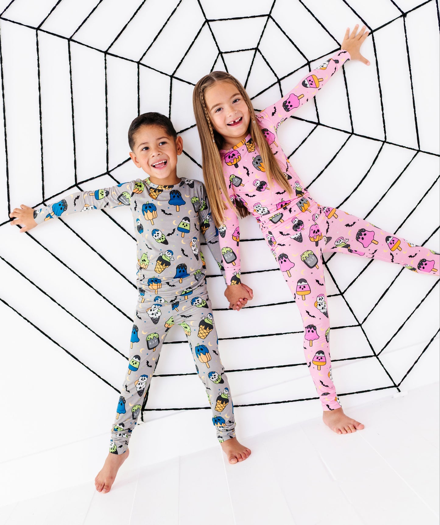 Two children wearing long-sleeve pajama sets with cartoon prints, lying on a white floor inside a large black spiderweb outline. The boy's pajamas are light gray with multicolored cartoon characters, and the girl's pajamas are pink with penguins.