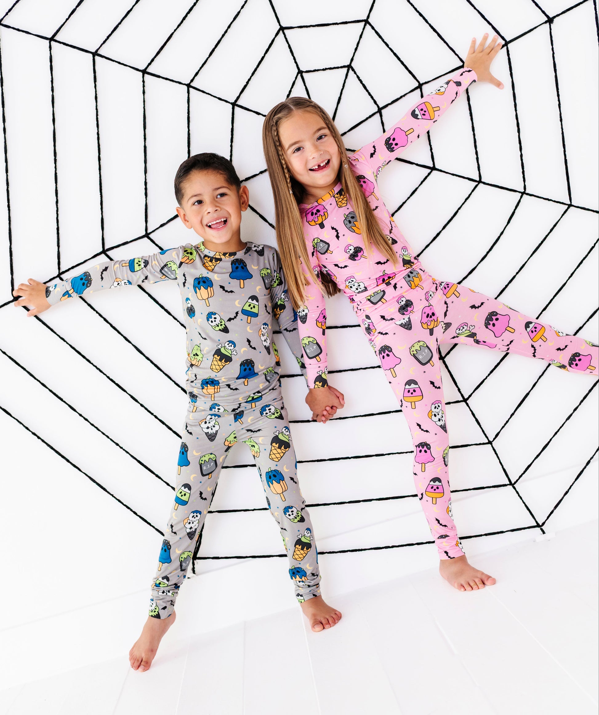 Two children wearing long-sleeve pajama sets with cartoon prints, lying on a white floor inside a large black spiderweb outline. The boy's pajamas are light gray with multicolored cartoon characters, and the girl's pajamas are pink with penguins.