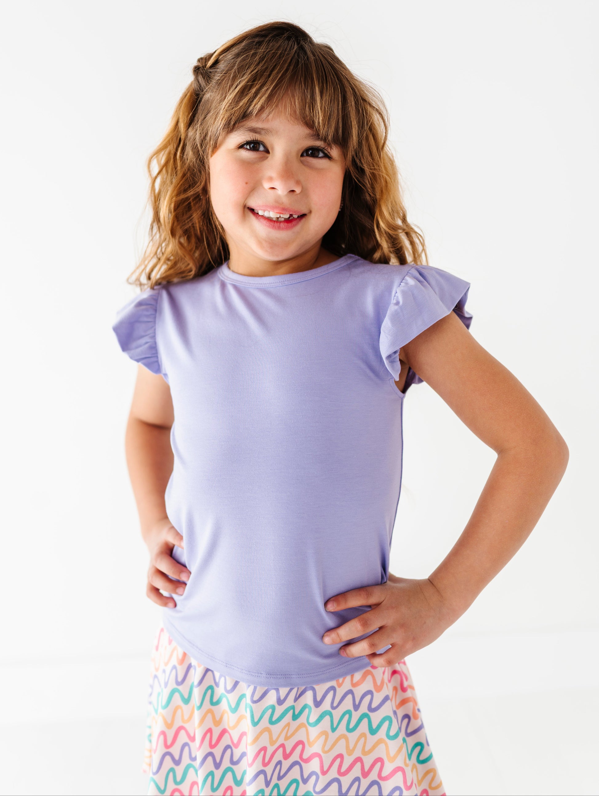 Young girl wearing a purple bamboo/cotton top with ruffled sleeves and a colorful skort on a white background