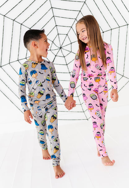Two children wearing matching long-sleeve pajamas with Halloween-themed prints, standing barefoot and holding hands in front of a large spiderweb backdrop. The boy's pajama set is light gray with colorful monster prints, and the girl's is pink with monster prints.