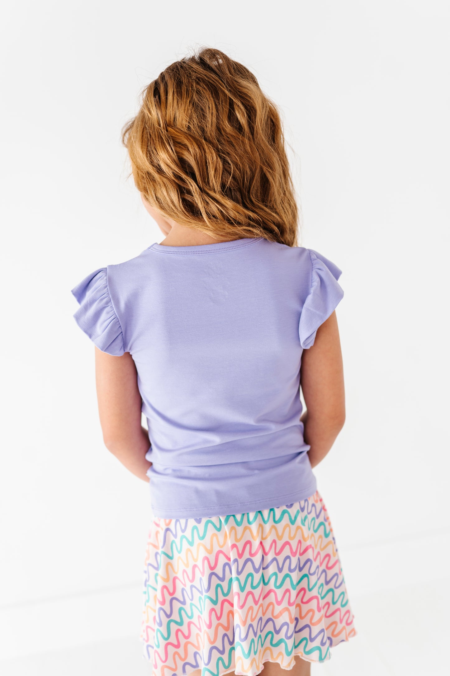 Young girl wearing a light purple bamboo/cotton top and colorful skort on a white background