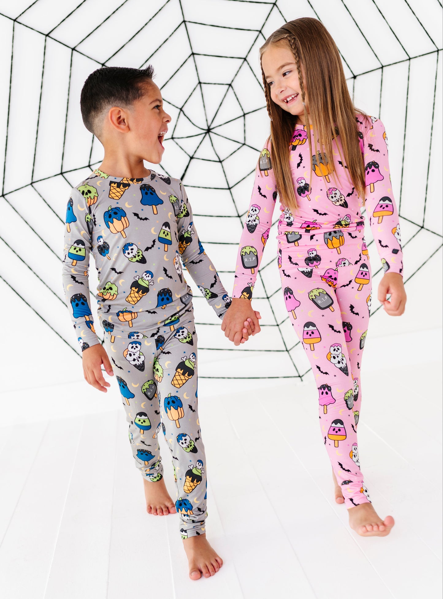 Two children wearing matching long-sleeve pajamas with Halloween-themed prints, standing barefoot and holding hands in front of a large spiderweb backdrop. The boy's pajama set is light gray with colorful monster prints, and the girl's is pink with monster prints.