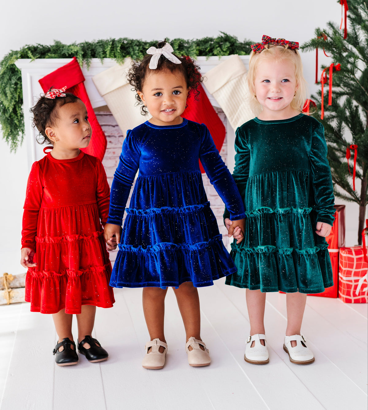 Three young children wearing matching velvet Christmas dresses in red, royal blue, and emerald green, standing together in front of a decorated holiday mantel with stockings and a Christmas tree.