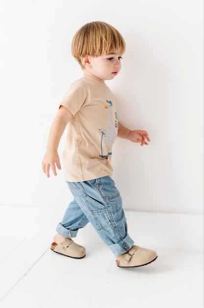 Young child wearing a beige t-shirt with a graphic and blue jeans, standing on a white floor.